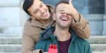 One man covers his partners eyes as he presents him with a gift wrapped with a red bow