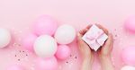 Womans hands holding gift or present box on pink pastel table decorated balloons and confetti. Flat lay style.