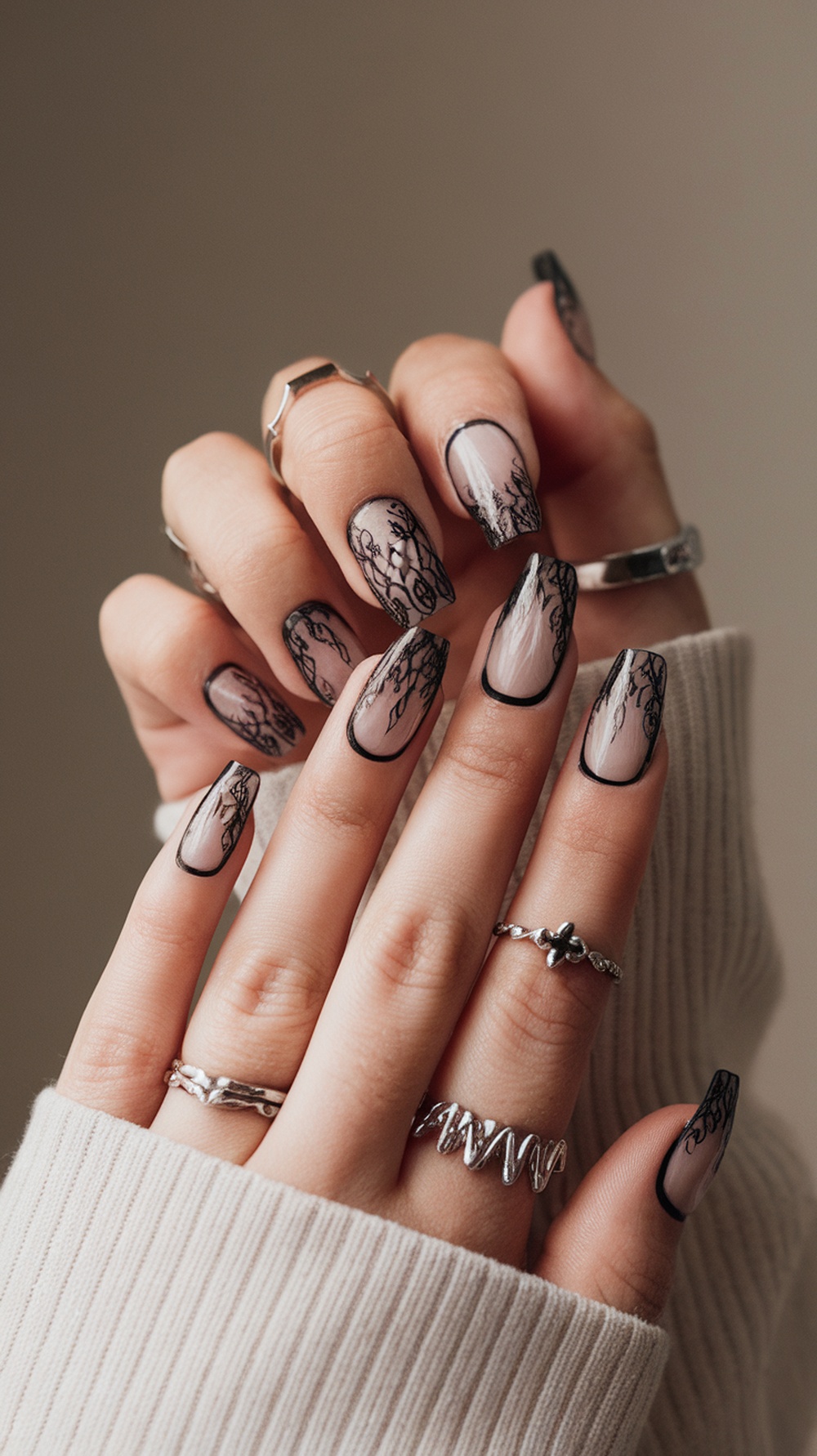 Close-up of hands with gothic glam nails featuring black lace designs on a nude base, adorned with silver rings.