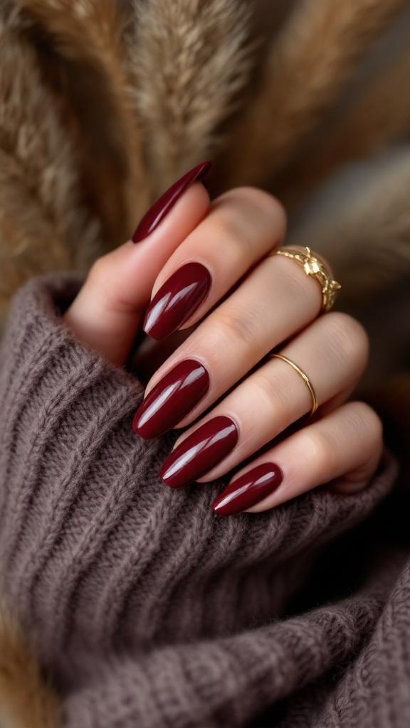 A hand with dark cherry red nails in an almond shape, wearing gold rings, resting on a cozy knit sweater.