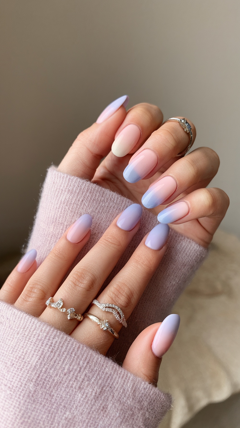 A hand showing pastel ombre nails transitioning from pink to blue, adorned with rings.