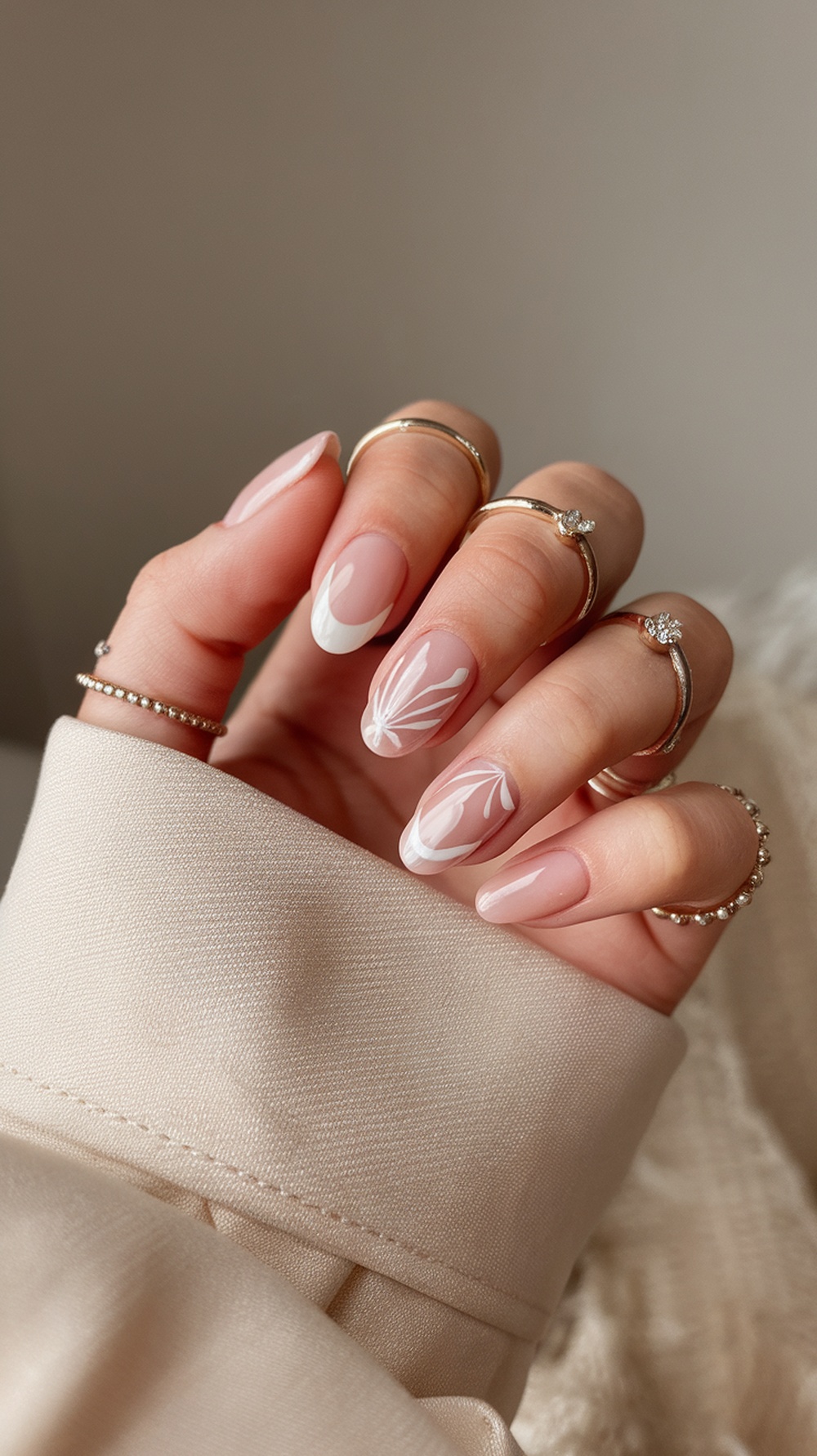 A hand with pastel nude nails featuring white floral designs, adorned with gold rings.